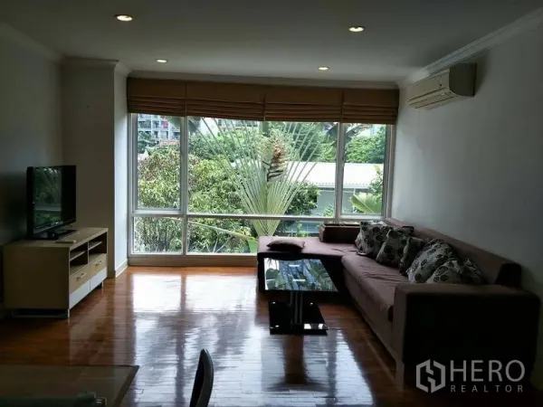 Condominium for rent in Khlong Toei, Bangkok - Bright living room with hardwood floors, L-shaped sofa, glass coffee table, TV console, and a large window with leafy garden view.