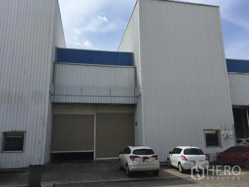 Factory for rent in Khlong Luang, Pathum Thani - Modern metal-clad factory facade with a large roller shutter door and cars parked on the driveway.