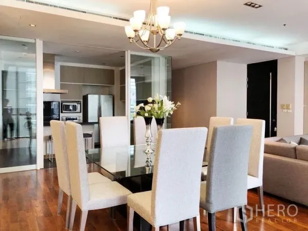 Condominium for rent in Khlong Toei, Bangkok - Bright dining room with glass table, upholstered chairs and chandelier beside glass partition to the kitchen.