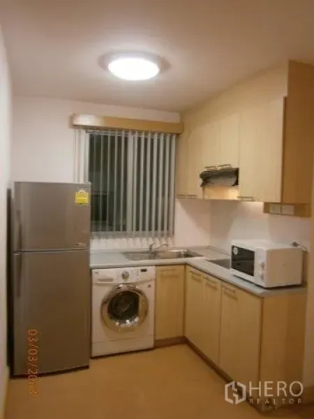 Condominium for rent in Khlong Toei, Bangkok - Compact kitchen with light wood cabinets, fridge, washing machine, microwave, and stainless sink.