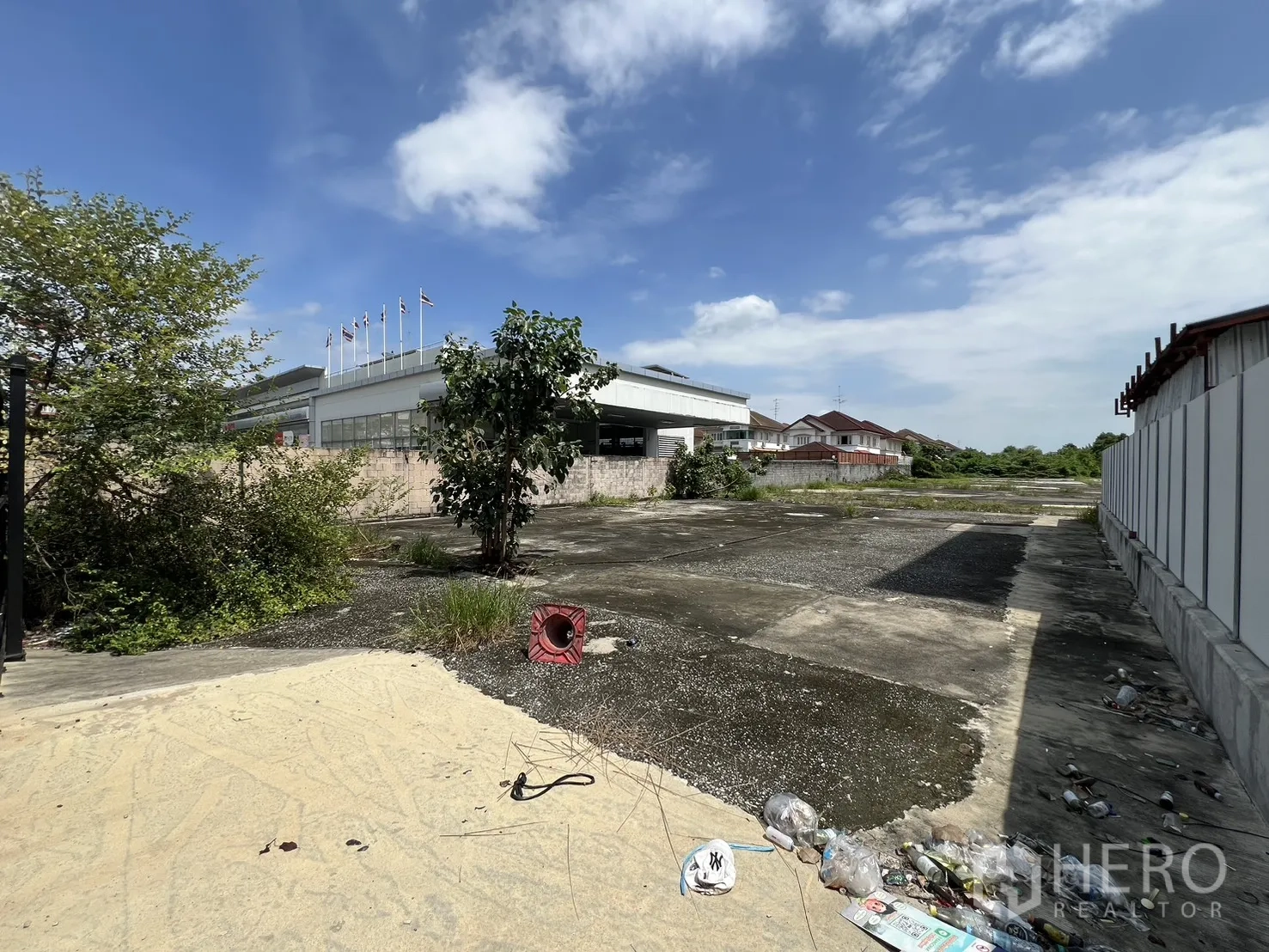 Land for sale in Bang Kapi, Bangkok - Edge of the land next to a tall commercial building and boundary wall, with sparse vegetation.