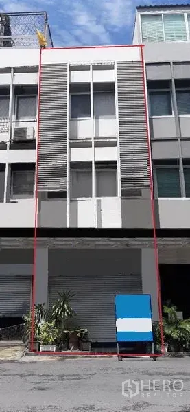 Shophouse for rent in Watthana, Bangkok - Modern multi-storey shophouse facade with a ground-floor roller shutter, vertical louver panels and potted plants, daytime street view in the Thonglor area.