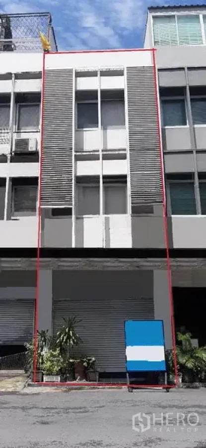 店屋 出租在Watthana, Bangkok - 现代多层店屋立面，底层卷帘门与竖向百叶，摆放盆栽的街景，通罗区域白天实景