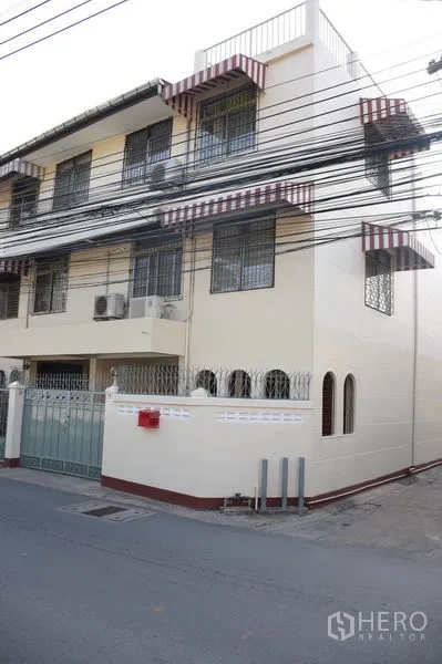 Townhouse for rent in Watthana, Bangkok - Corner view of cream townhouse with high wall, barred windows and quiet street.
