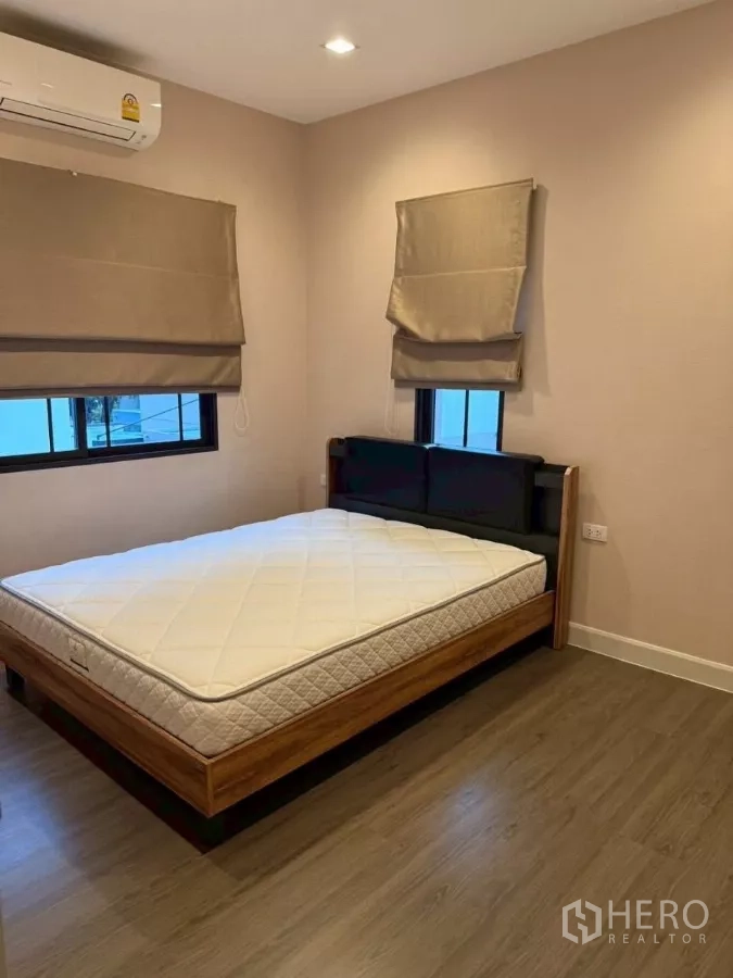 Detached House for rent or sale in Bang Phli, Samut Prakan - Secondary bedroom with double bed, corner windows, wall‑mounted air conditioner and wood‑look floors.