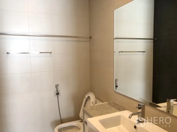 Condominium for rent or sale in Khlong Toei, Bangkok - Modern bathroom with large mirror, vanity sink, toilet and handheld bidet on neutral tiles.