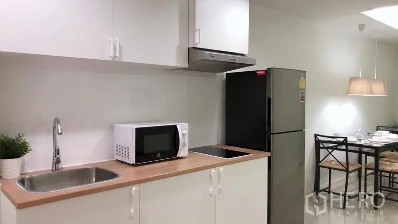 Condominium for rent or sale in Khlong Toei, Bangkok - White kitchenette with sink, cooktop, hood, microwave, and stainless refrigerator next to dining set.