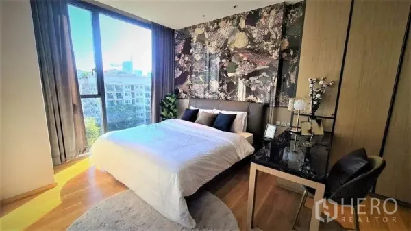 Condominium for rent in Khlong Toei, Bangkok - Bedroom with large window, city view, statement wall behind the bed and a work desk.