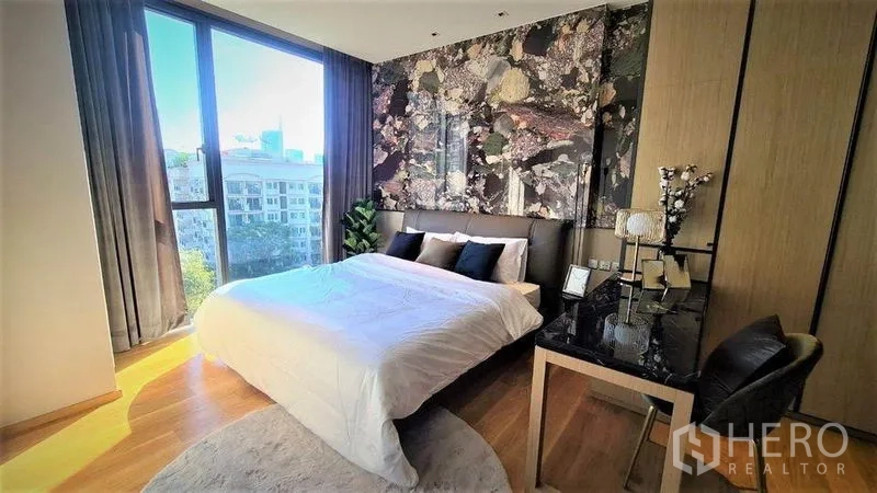 Condominium for rent in Khlong Toei, Bangkok - Bedroom with large window, city view, statement wall behind the bed and a work desk.