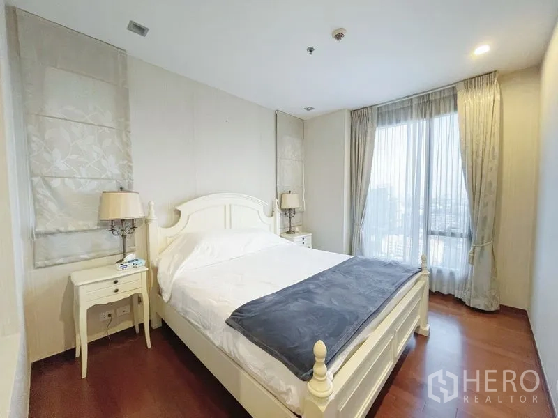 Condominium for sale in Khlong Toei, Bangkok - Master bedroom with white bed, bedside lamps and a large curtained window for natural light.