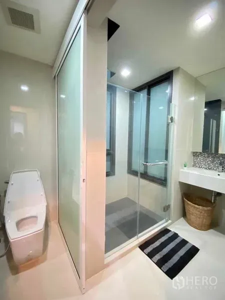 Condominium for rent in Khlong Toei, Bangkok - Modern bathroom featuring a glass-enclosed shower, frosted partition, toilet, vanity and window.