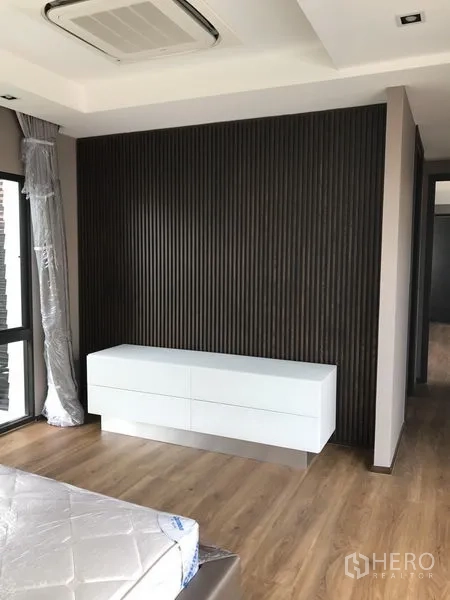 Detached House for rent in Bang Kapi, Bangkok - Bedroom corner with dark slatted feature wall and white low cabinet on wood floors.