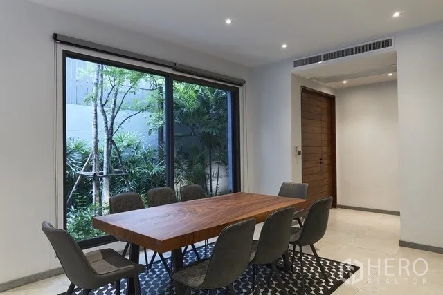 Townhouse for sale in Watthana, Bangkok - Dining zone with a solid wood table beside a picture window framing trees.