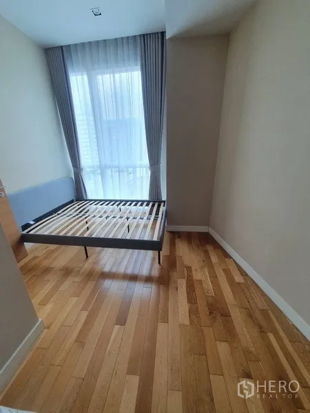 Condominium for rent in Khlong Toei, Bangkok - Cozy second bedroom with bed frame, large window, and light wood floor.