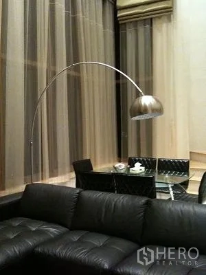 Condominium for rent or sale in Khlong Toei, Bangkok - Double-height living room with black leather sofa, arc floor lamp, and sheer curtains.