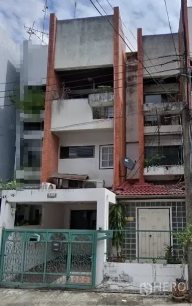 Shophouse for sale in Khlong Toei, Bangkok - Street-facing multi-storey shophouse in Bangkok with red brick columns, balconies with plants, a green metal gate, gated carport, and overhead utility lines.
