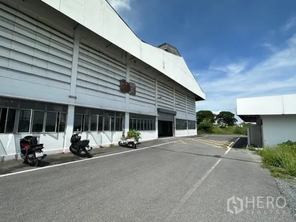 Warehouse for rent in Lam Luk Ka, Pathum Thani - Large white warehouse exterior with loading bay, scooters parked along the wall, and a wide internal driveway under a clear sky.