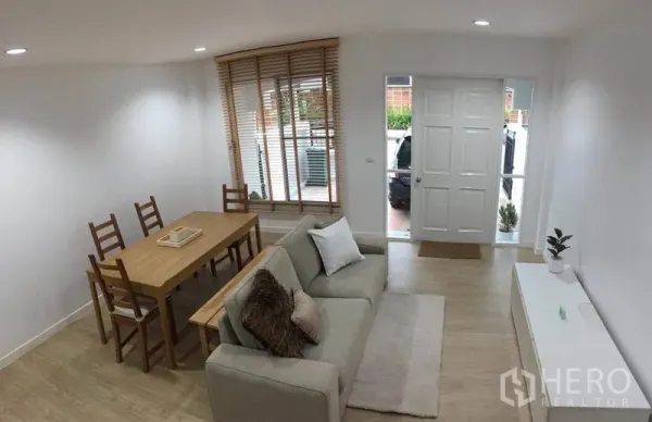 Townhouse for rent in Lat Phrao, Bangkok - Living room view to the entrance showing sofa, dining set and window blinds.