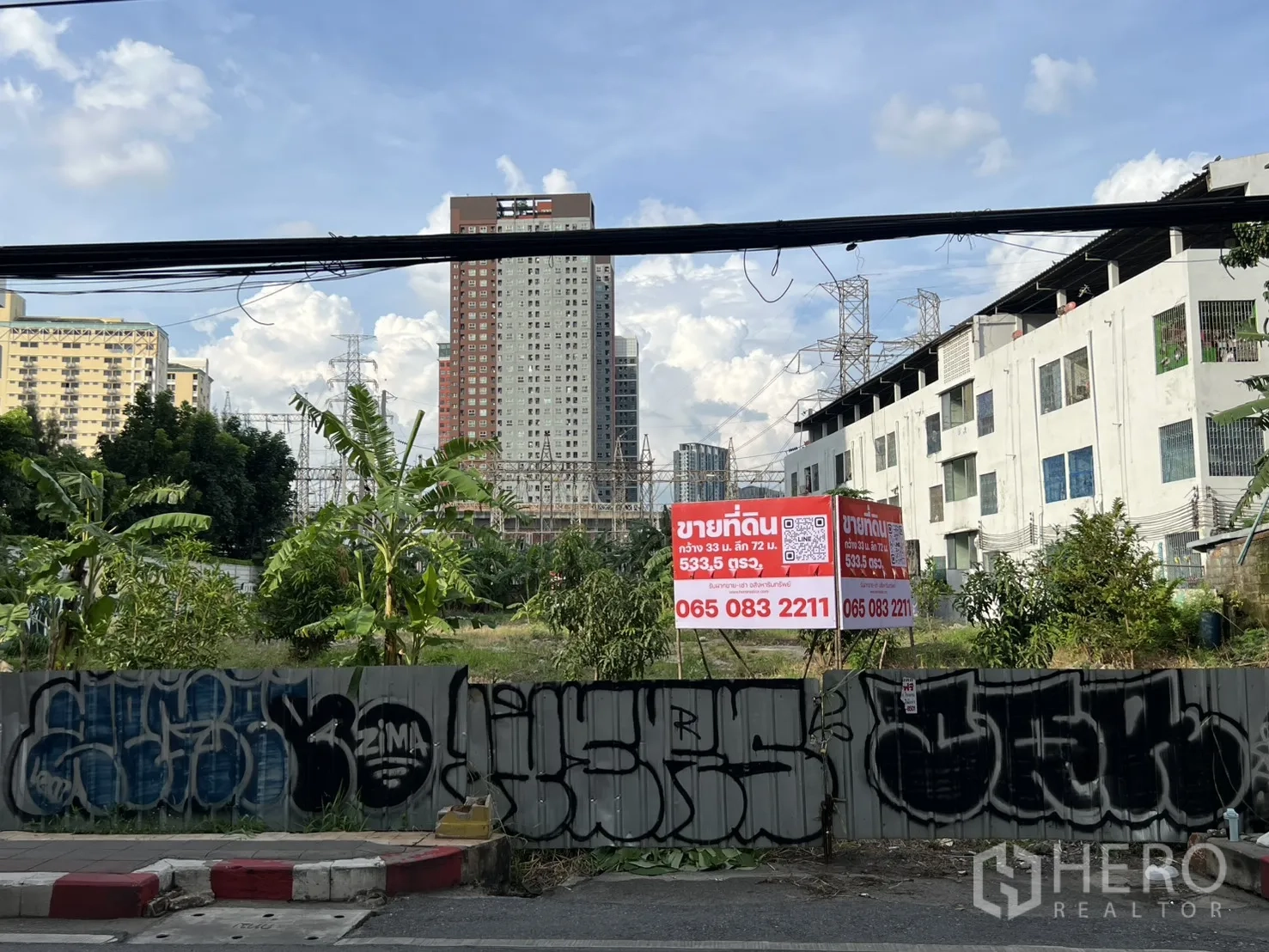 Land for sale in Suan Luang, Bangkok - Frontage view from the road with a fence, graffiti, and a sale sign leading to the open plot and skyline.