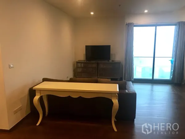 Condominium for rent in Khlong Toei, Bangkok - Living room with wood floors, sofa, TV console and sliding glass doors to a balcony.
