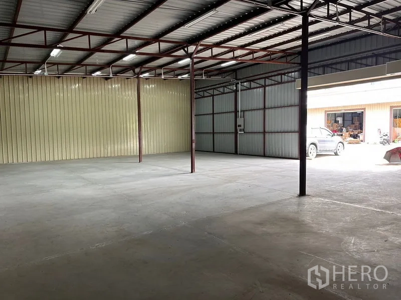 Warehouse for rent in Mueang Nonthaburi, Nonthaburi - Open-plan warehouse showing concrete slab, steel columns, and daylight from the loading door with parking outside.
