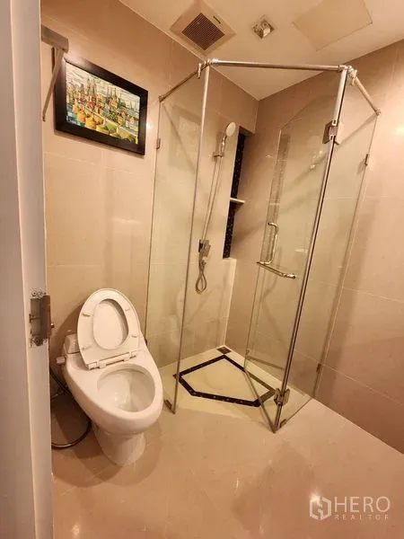 Condominium for rent in Bang Rak, Bangkok - Beige bathroom featuring a glass-enclosed shower, handheld spray and toilet with wall artwork.