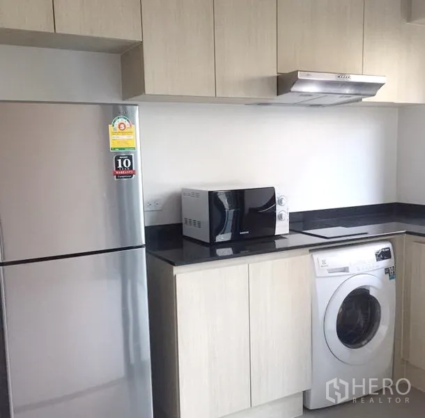 Townhouse for rent in Suan Luang, Bangkok - Fitted kitchen with light wood cabinets, black countertop, stainless fridge, microwave and front‑load washer.