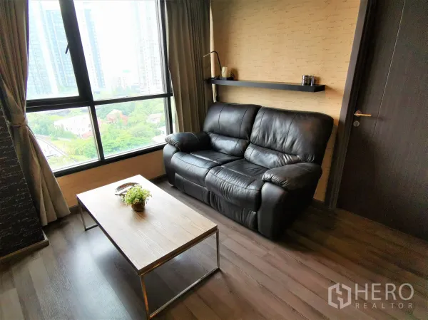 Condominium for rent or sale in Watthana, Bangkok - Sofa corner and wooden coffee table beside a floor-to-ceiling window overlooking greenery and towers.