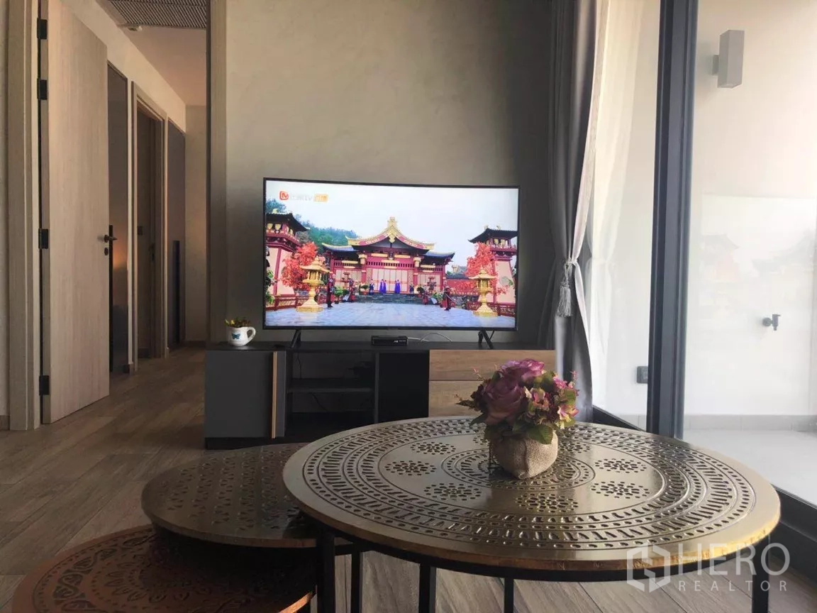 Condominium for sale in Watthana, Bangkok - TV area with a large flat-screen on a console, round metal coffee tables with a small flower vase, and sliding glass doors to the balcony.