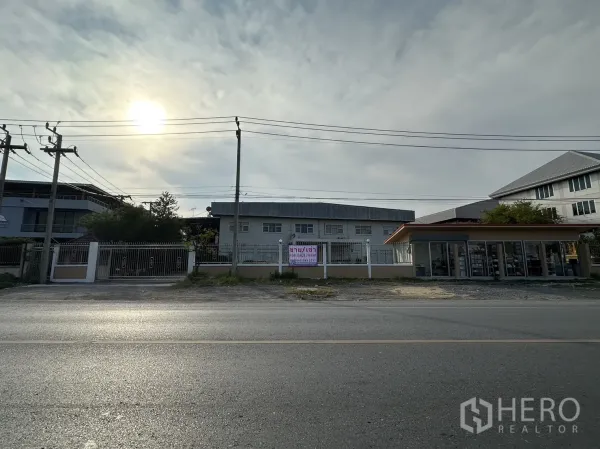 Factory for rent or sale in Lam Luk Ka, Pathum Thani - Wide roadside view of the factory frontage with a for sale/rent banner and a neighboring shop.