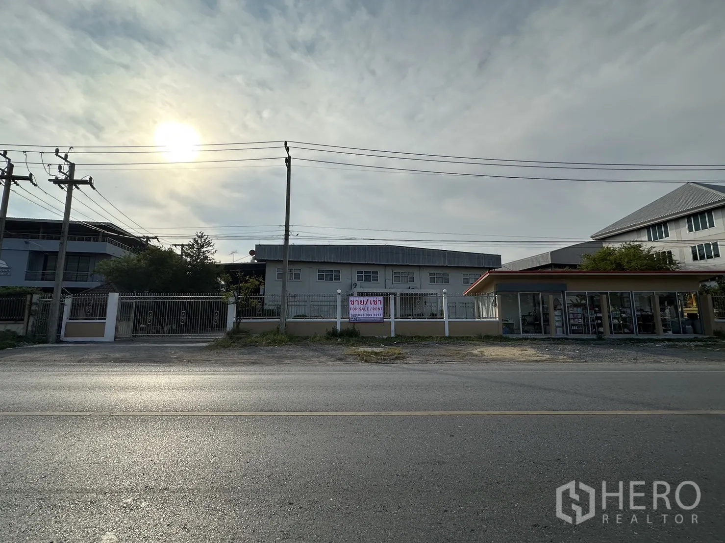 Factory for rent or sale in Lam Luk Ka, Pathum Thani - Wide roadside view of the factory frontage with a for sale/rent banner and a neighboring shop.