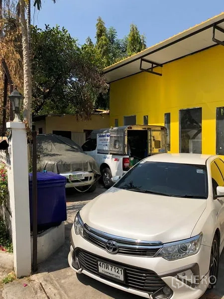 独立式住宅 出售在Khlong Toei, Bangkok - 房前车道可停车，多辆车停靠在亮黄色外立面旁并有雨篷遮阳。
