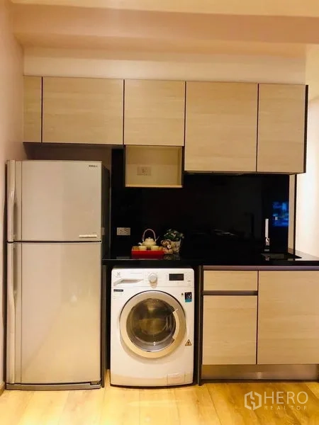 Condominium for rent in Khlong Toei, Bangkok - Compact modern kitchen with light wood cabinets, refrigerator, black backsplash and a front-load washing machine.