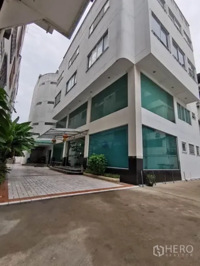 办公空间 出租在Khlong San, Bangkok - 多层办公楼转角外观，绿色玻璃窗与有顶门廊车道。