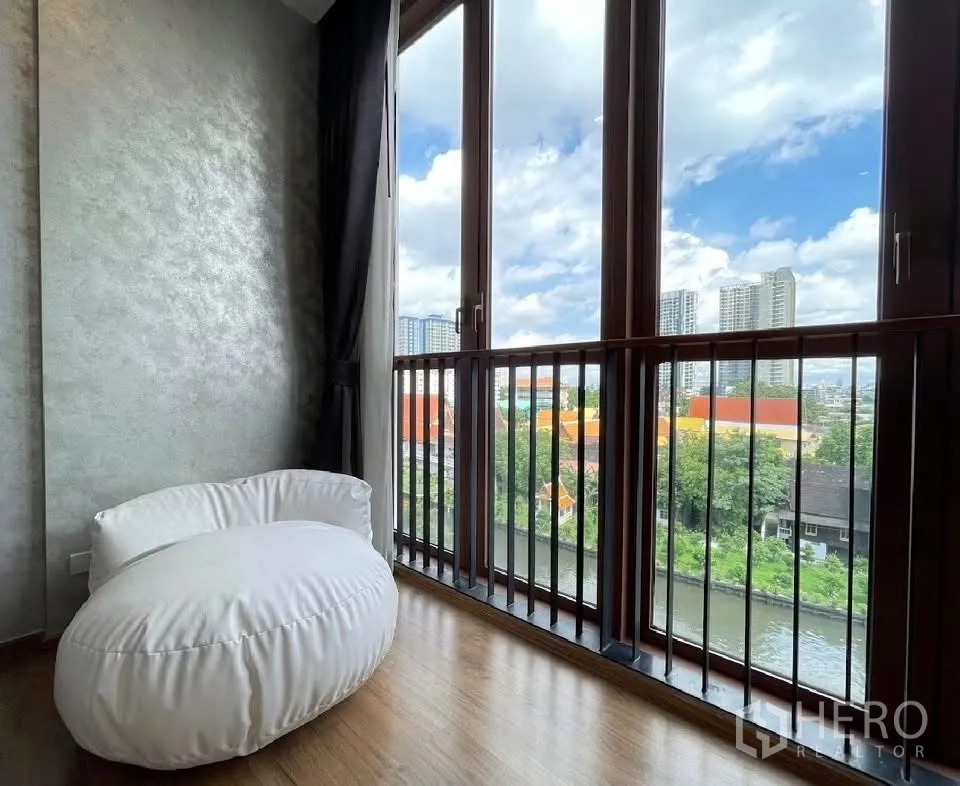 Condominium for rent in Watthana, Bangkok - Cozy corner with a white beanbag by tall windows overlooking a canal and skyline.