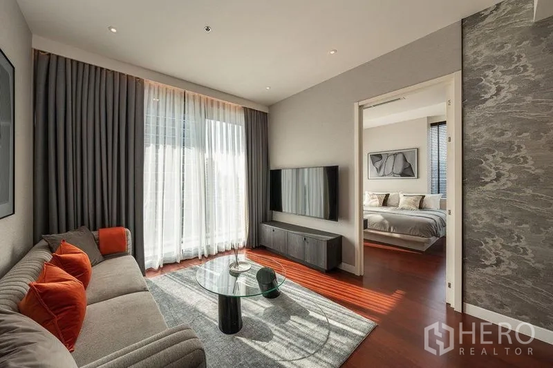 Condominium for rent in Watthana, Bangkok - Living room with floor-to-ceiling curtains, wall-mounted TV console, and doorway leading to a bedroom.