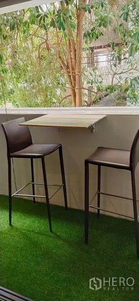 Condominium for sale in Watthana, Bangkok - Balcony with green turf flooring, foldable wall table and two high stools overlooking leafy trees.