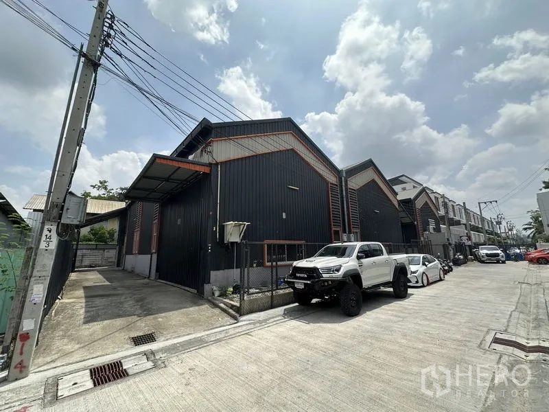 Warehouse for rent in Lat Phrao, Bangkok - Street view of a black metal-clad warehouse with gable roof and gated parking along a quiet lane in Lat Phrao.
