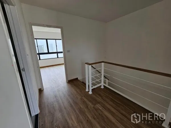 Townhouse for rent in Bang Kapi, Bangkok - Upper-floor landing with wood-look flooring and a white metal staircase leading to a bright bedroom with large windows.