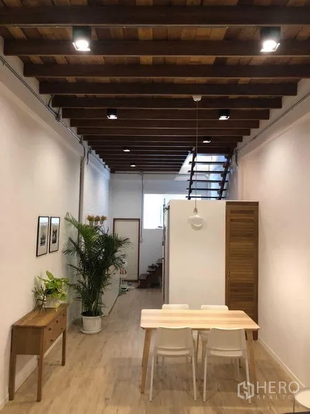 Detached House for rent in Phra Nakhon, Bangkok - Dining space under exposed wooden beams with a light wood table and staircase in the background.