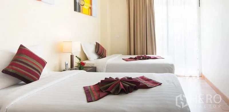 Condominium for rent in Khlong Toei, Bangkok - Twin bedroom with crisp white linens, accent pillows and a bedside lamp, filled with natural light through full-height curtains.