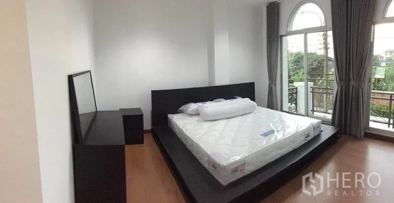 Townhouse for rent in Lat Phrao, Bangkok - Spacious master bedroom with king bed, arched window, balcony doors and curtains.