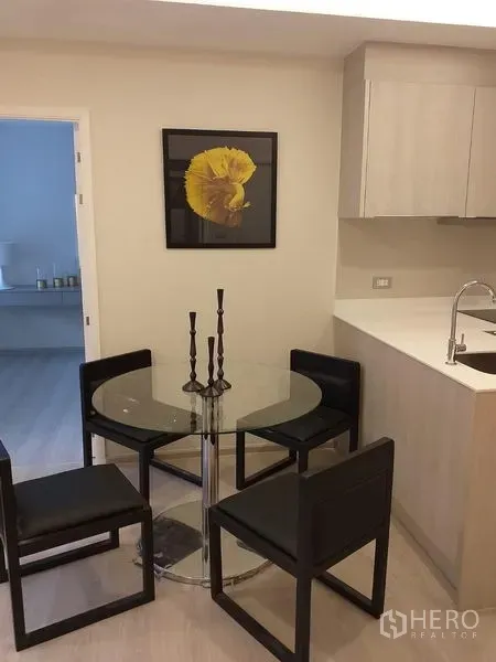 Condominium for rent in Khlong Toei, Bangkok - Glass dining table with four black chairs beside a light-wood kitchenette and yellow artwork.