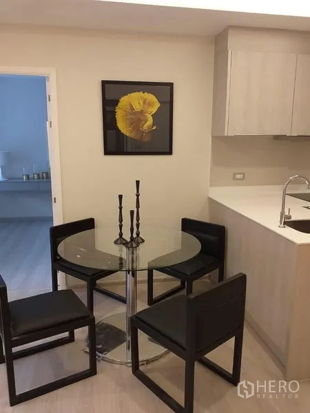 Condominium for rent in Khlong Toei, Bangkok - Glass dining table with four black chairs beside a light-wood kitchenette and yellow artwork.