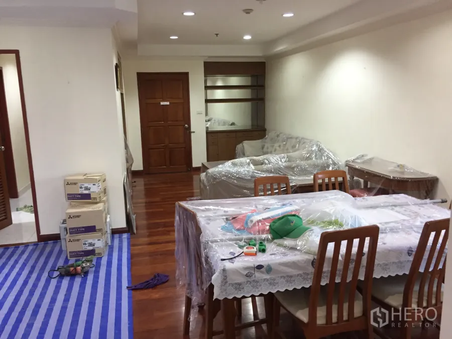 Condominium for rent in Watthana, Bangkok - Open-plan living and dining room with hardwood floors, recessed lighting, built-in shelving, and furniture wrapped for move-in.