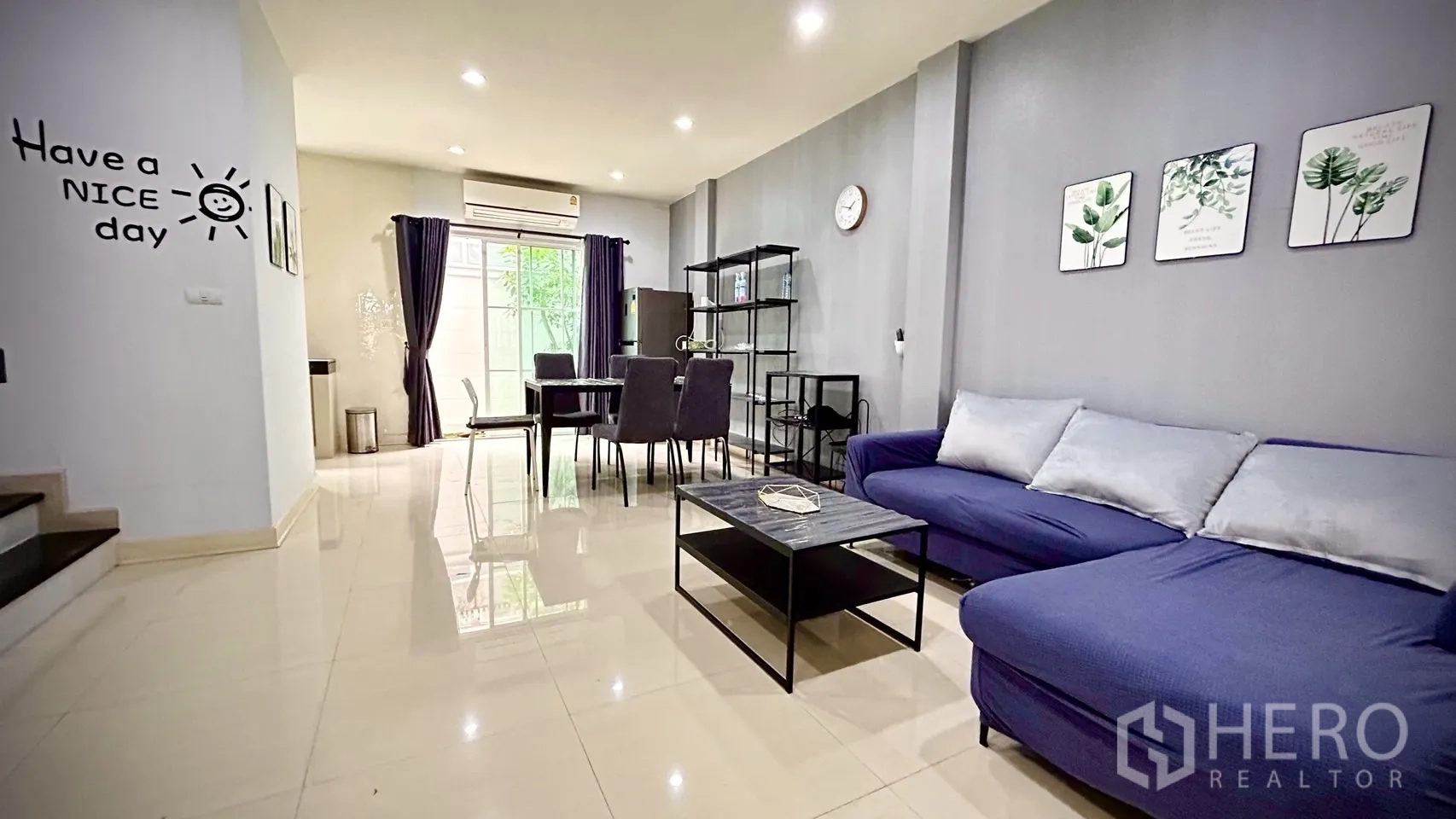 Townhouse for rent in Bang Kapi, Bangkok - Open-plan living and dining area with blue sectional sofa, dining table and sliding glass door to a patio.