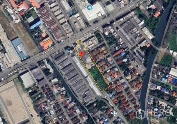 Land for sale in Bang Kapi, Bangkok - Entrance area of the cleared plot beside an industrial building and trees, flags visible on the roofline.