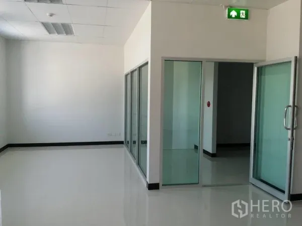 Warehouse for rent in Samut Prakan - Glass-partitioned office corridor with sliding doors and an illuminated emergency exit sign.