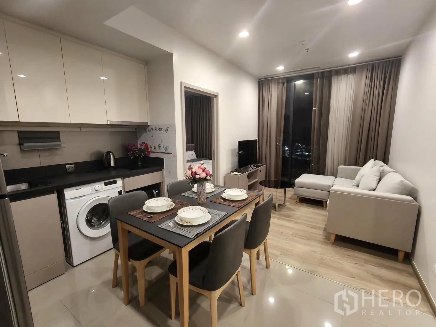 Condominium for rent in Khlong Toei, Bangkok - Open-plan living and dining area with L-shaped sofa, TV, and a kitchen with washing machine, plus a night city view through tall windows.