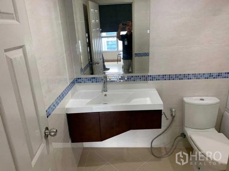 Condominium for rent in Bang Rak, Bangkok - Modern bathroom with large mirror, white vanity, blue mosaic trim and toilet.